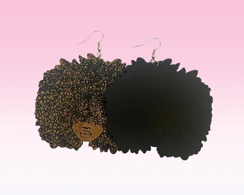 May include: A pair of wooden earrings featuring a black silhouette of a woman with an afro hairstyle. One earring has a detailed design of the woman's face and hair, while the other is a solid black afro.