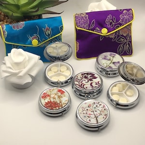 Puede incluir: Seis cajas redondas para pastillas con diferentes diseños florales. Las cajas para pastillas son plateadas con fondo blanco y tienen una tapa con bisagras. Las cajas para pastillas están dispuestas en círculo sobre una superficie blanca. También hay dos pequeñas bolsas de satén con diseños florales en la imagen. Una bolsa es azul y la otra es morada.
