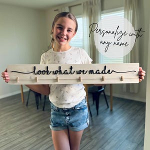 May include: A wooden sign with the words "look what we made" in black script. The sign has five small wooden clips and is held by a person. The sign is light-colored with a distressed finish. The text "Personalize with any name" is in the background.