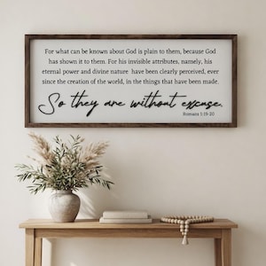 May include: A framed wooden sign with the text "For what can be known about God is plain to them... So they are without excuse." A wooden table holds a vase of dried flowers, books, and a beaded garland. The sign is mounted on a white wall.