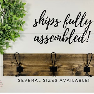 May include: A wooden wall-mounted coat rack with three black metal hooks. The dark brown stained wood is mounted on a white background. Text reads "ships fully assembled!" and "SEVERAL SIZES AVAILABLE!"