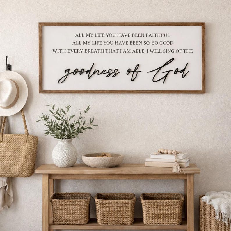 May include: A wooden framed sign with the words "ALL MY LIFE YOU HAVE BEEN FAITHFUL" and "goodness of God" in black script. The sign hangs above a wooden console table with woven baskets and a vase of greenery.