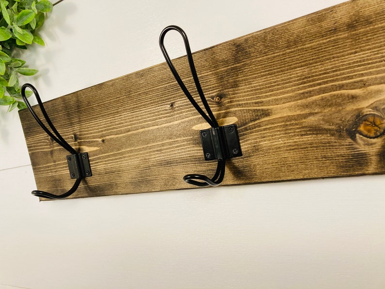 May include: A rustic wooden coat rack with two black metal hooks. The wood is stained a dark brown, showcasing the natural grain. The hooks are mounted on black metal plates, creating a contrast against the wood. Perfect for entryways or bedrooms.