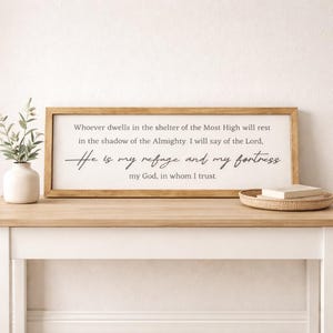 May include: A rectangular wooden framed sign with the text "Whoever dwells in the shelter of the Most High will rest in the shadow of the Almighty. He is my refuge and my fortress". The sign is on a light wood surface.