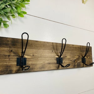 May include: A rustic wooden coat rack with three black metal hooks. The wood is stained a dark brown, and the hooks are mounted on black metal plates. The rack is designed for hanging coats, hats, or other items.