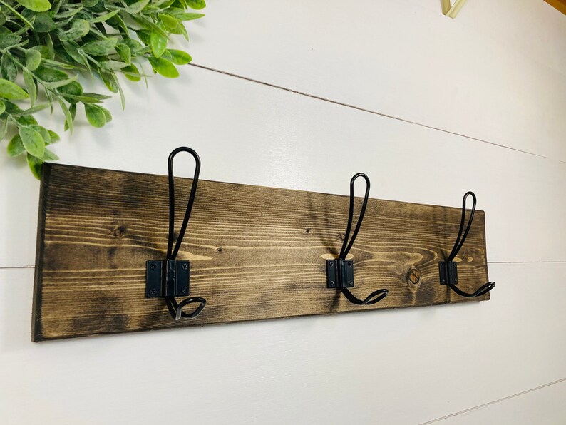 May include: A rustic wooden coat rack with three black metal hooks. The wood is stained a dark brown, and the hooks are mounted on black metal plates. The rack is designed for hanging coats, hats, or other items.