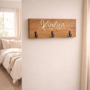 May include: A wooden key holder with the name "Kaelyn" in a cursive font. The key holder is made of a light brown wood and has three black metal hooks. The key holder is mounted on a white wall.