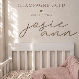 May include: A wall-mounted sign featuring the name "josie ann" in a cursive script font, positioned above a white crib with a floral patterned sheet and a dusty pink muslin blanket. The text "CHAMPAGNE GOLD" and "COLOR OPTION" appear above.