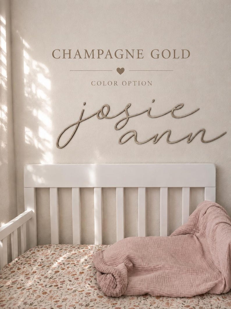 May include: A wall-mounted sign featuring the name "josie ann" in a cursive script font, positioned above a white crib with a floral patterned sheet and a dusty pink muslin blanket. The text "CHAMPAGNE GOLD" and "COLOR OPTION" appear above.