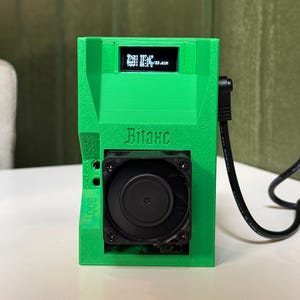 May include: A bright green electronic device with a small screen displaying data. The device has a black fan, a power cable, and the word "Bitaxe" printed on the front. The device is on a white surface.