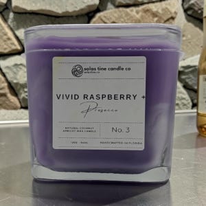 May include: A square glass candle filled with purple wax. The white label reads Solas Tine Candle Co, Vivid Raspberry + Prosecco, No. 3, Natural Coconut Apricot Wax Candle, 12oz - 340g, and Handcrafted in Florida.