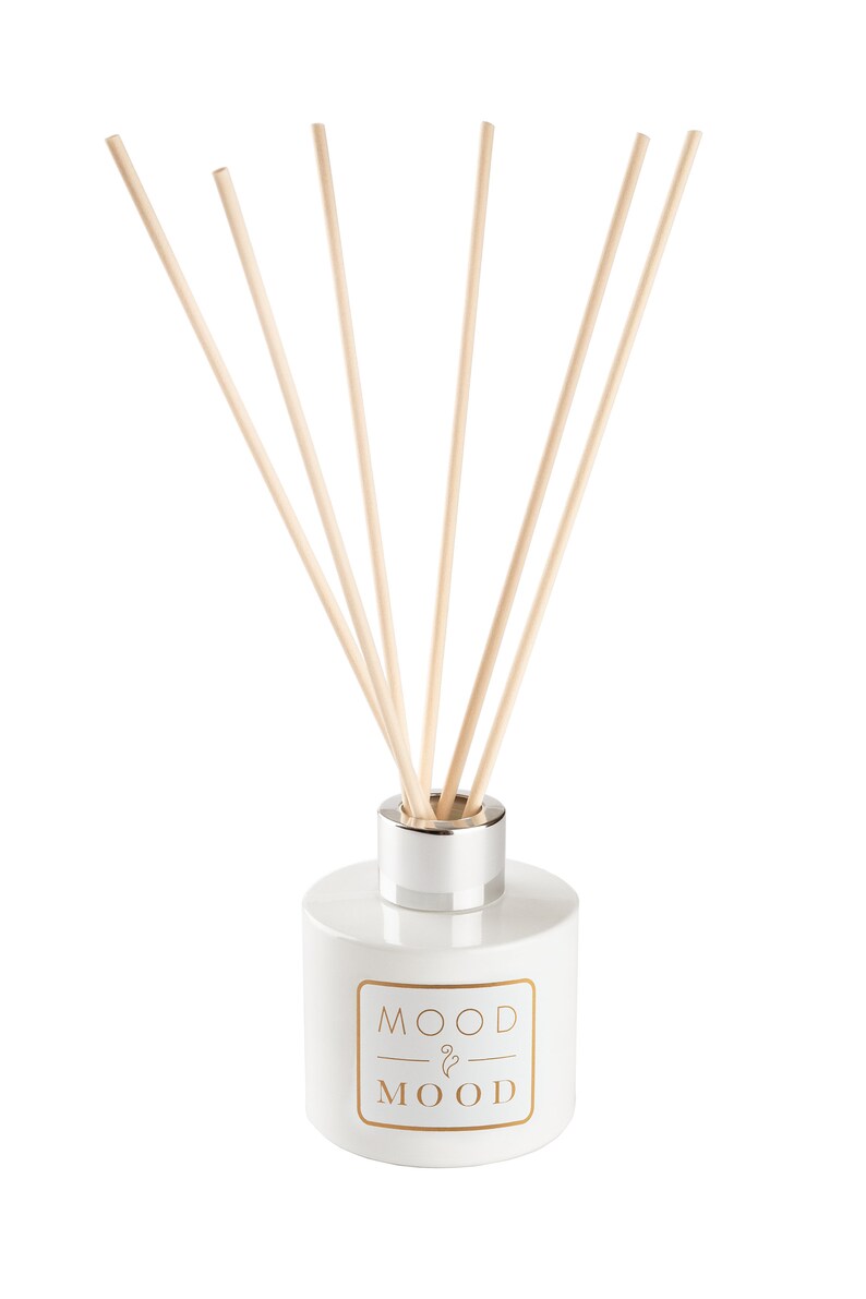 Op de afbeelding: Witte keramische diffuserfles met een zilveren deksel en houten stokjes. De fles heeft de tekst "MOOD & MOOD" in goud gedrukt.