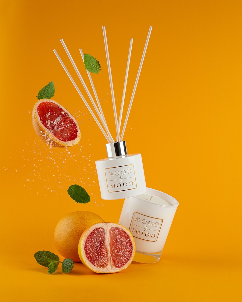 Op de afbeelding: Twee witte flessen met de tekst "MOOD & MOOD" erop. De ene fles is een diffuser met stokjes en de andere is een kaars. De flessen zijn omringd door grapefruitpartjes en groene bladeren op een gele achtergrond.