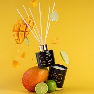 May include: A black diffuser bottle and candle with the text "MOOD & MOOD" on the front. The diffuser bottle has white sticks and the candle is surrounded by mango slices, lime slices, and a yellow background.
