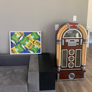 Może przedstawiać: Jukebox w stylu vintage w czerwono-żółto-brązowej kolorystyce. Jukebox posiada gramofon, radio i odtwarzacz CD. Jukebox stoi przed szarą kanapą i oprawionym wydrukiem w stylu pop art z puszkami zupy Campbell.