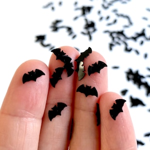 May include: Black bat shaped confetti, scattered on a white surface. A hand is holding a handful of the confetti.