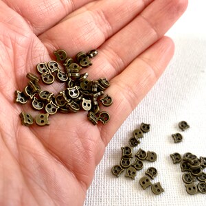 May include: A collection of small, bronze-colored metal buttons shaped like cat faces. The buttons have two holes for sewing and are scattered across a surface. The cat face buttons are a craft supply.