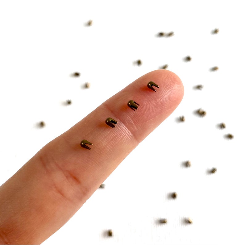 May include: A close-up of a finger holding four small, gold-colored metal rivets. The rivets have a rounded top and a pointed bottom. There are many more rivets scattered on a white surface.