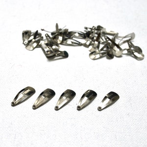 Op de afbeelding: Een verzameling zilverkleurige metalen haarspeldjes. Sommige zijn verspreid, andere netjes gerangschikt. De haarspeldjes zijn ongeveer 2,5 cm lang en hebben een eenvoudig, functioneel ontwerp. Het metaal heeft een glanzend oppervlak.