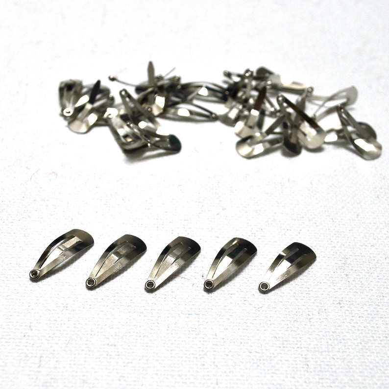 Op de afbeelding: Een verzameling zilverkleurige metalen haarspeldjes. Sommige zijn verspreid, andere netjes gerangschikt. De haarspeldjes zijn ongeveer 2,5 cm lang en hebben een eenvoudig, functioneel ontwerp. Het metaal heeft een glanzend oppervlak.