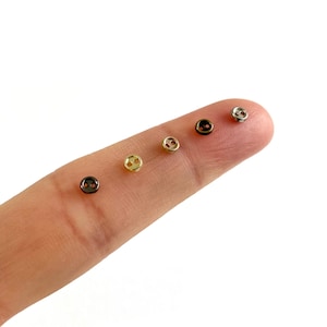 Könnte beinhalten: Eine Sammlung kleiner, runder Knöpfe in verschiedenen metallischen Ausführungen, darunter Gold, Silber und Schwarz. Jeder Knopf hat zwei Löcher zum Annähen. Die Knöpfe werden auf einem Finger vor weißem Hintergrund gezeigt.