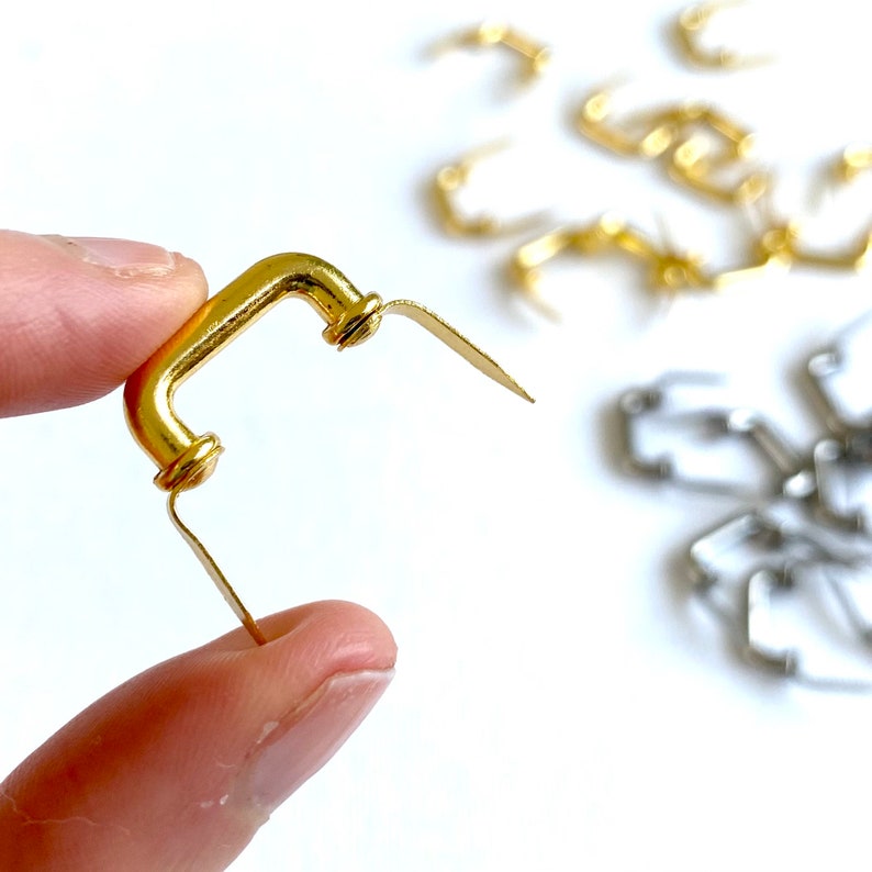 K&ouml;nnte beinhalten: Goldfarbener Metallgriff mit abgerundeter rechteckiger Form und einer flachen, abgewinkelten R&uuml;ckplatte. Der Griff wird in einer Hand gehalten, und im Hintergrund befinden sich weitere &auml;hnliche Griffe.
