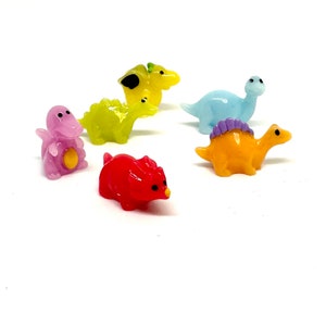 May include: A collection of six miniature dinosaur figurines in various colors. The dinosaurs include a purple triceratops, a red stegosaurus, a yellow brontosaurus, a light blue apatosaurus, a green stegosaurus, and an orange triceratops.