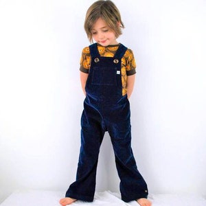 Könnte beinhalten: Ein Kind trägt eine marineblaue Cord-Latzhose mit verstellbaren Trägern und Knopfverschluss. Darunter ist ein kurzärmeliges Hemd mit einem braunen und gelben Muster zu sehen. Geeignet für Kinderbekleidung.