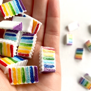 May include: A hand holding a pile of rainbow layered cake charms. The charms are made of white plastic with rainbow stripes. There are several charms scattered on a white surface.