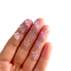 May include: A selection of small, translucent jellyfish charms in lavender and purple hues. Each charm has a rounded bell and trailing tentacles. The charms have a glittery, iridescent finish, adding sparkle. These decorative charms are ideal for crafting and jewellery making.
