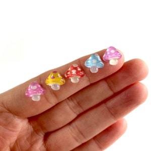 May include: Six colorful mushroom-shaped resin cabochons with white polka dots. The cabochons are arranged in a row on a hand, from left to right: pink, yellow, red, blue, purple.