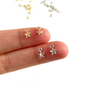 May include: Four gold and silver star-shaped charms with jump rings. The charms are on a finger.
