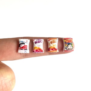May include: Four miniature bags of Japanese snacks. The bags are white with colorful Japanese characters and images of fruit and a cartoon monkey.