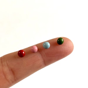 Op de afbeelding: Vier kleine ronde kralen in verschillende kleuren: rood, roze, lichtblauw en groen. De kralen liggen op een vinger.