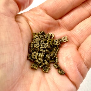 May include: A collection of small, antique brass-colored skull-shaped charms. The charms are detailed with eye sockets and a nose, and are scattered across a hand. These charms are likely used for jewelry making or other craft projects.