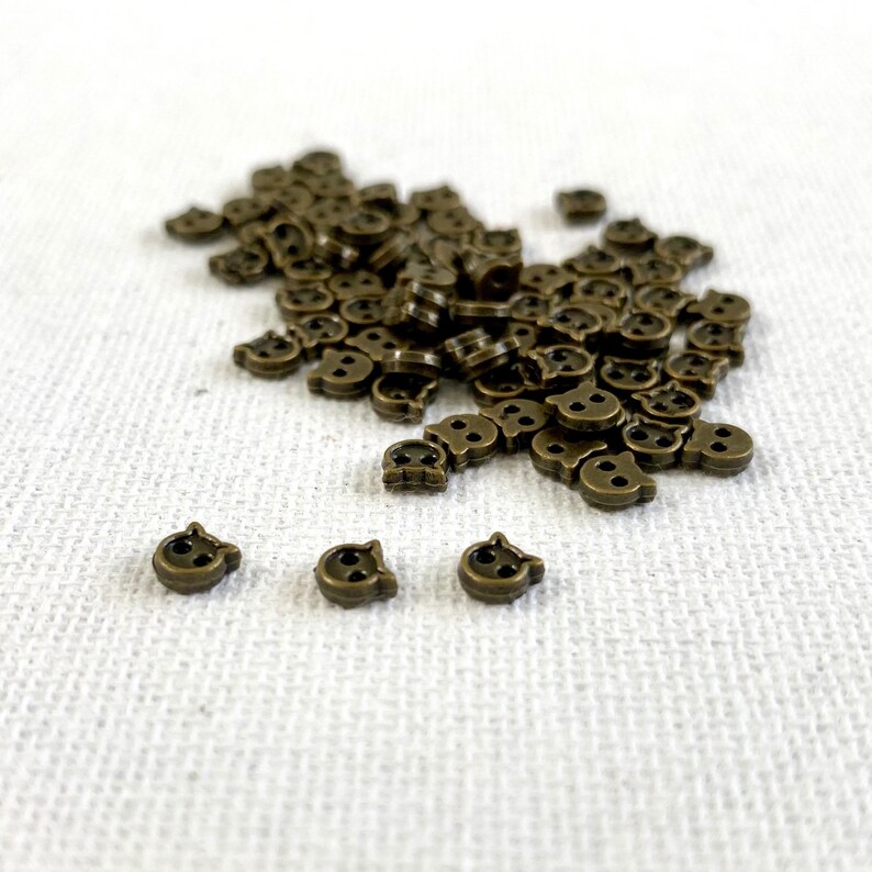 May include: A collection of small, bronze-colored skull-shaped buttons scattered on a white surface. Each button has two eye holes and a nose, with two small holes for sewing. The buttons are likely for crafts or clothing.