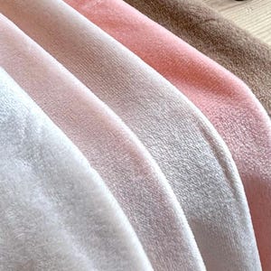 May include: Close-up of soft, plush fabric in shades of white, light pink, medium pink, and brown. The fabric appears to be velvet or a similar material, with a subtle sheen. The image showcases the texture and color variations.