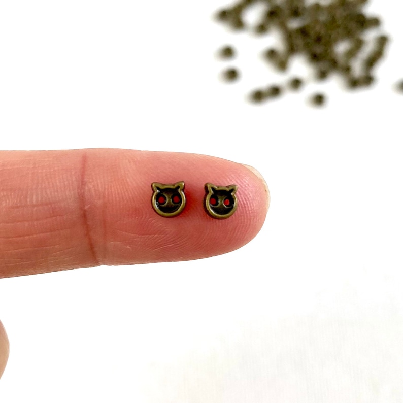 May include: Two small, antique bronze-colored cat-shaped charms with red eyes are displayed. The charms are held between a finger and thumb, with additional charms scattered in the background. These charms are ideal for jewelry making.
