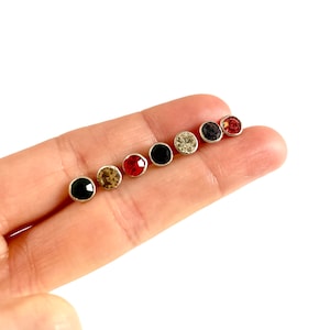 May include: A set of six small stud earrings with round, colorful rhinestones. The earrings are arranged in a row on a finger, with the rhinestones facing the viewer. The colors of the rhinestones are black, white, red, and brown.