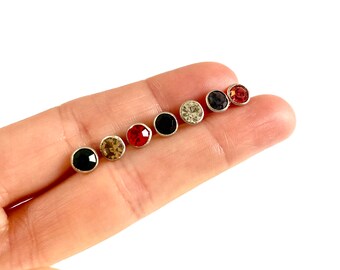 May include: A set of six small stud earrings with round, colorful rhinestones. The earrings are arranged in a row on a finger, with the rhinestones facing the viewer. The colors of the rhinestones are black, white, red, and brown.