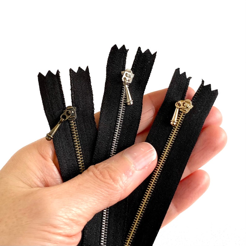 May include: Three black zipper tapes with different colored metal zipper pulls. The zipper pulls are silver, gold, and bronze.