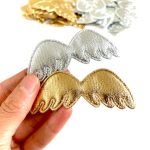 May include: Two pairs of angel wings, one pair is gold and the other is silver. The wings are made of a soft, textured fabric and have a slightly raised design.