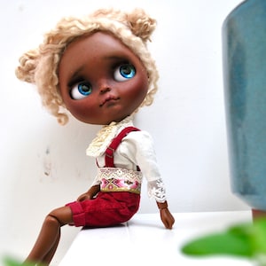 May include: A doll with blonde curly hair, brown skin, and blue eyes is wearing a white blouse with lace trim, red overalls, and a patterned belt. The doll is sitting on a white surface.