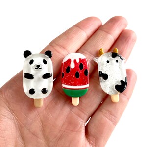 May include: Three miniature popsicle charms, each featuring a different design: a panda, a watermelon, and a cow. The charms are made of clear resin with glitter accents and are approximately 1 inch tall.