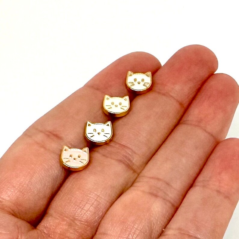 May include: Four gold-colored cat face charms with white enamel. The charms are arranged in a row on a hand.
