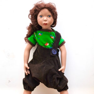 May include: A doll with brown hair and brown eyes wearing a green shirt with a bird pattern and brown overalls with a blue button.