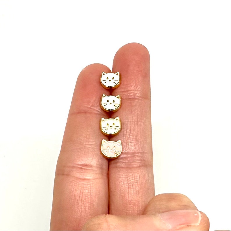 May include: Four gold-colored cat face charms with white enamel. The charms are lined up vertically on a finger.