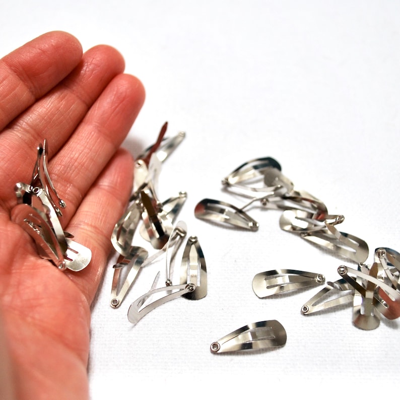 Op de afbeelding: Een verzameling zilverkleurige metalen haarspeldjes. De haarspeldjes zijn klein en hebben een eenvoudig, functioneel ontwerp. Sommige worden in een hand gehouden, terwijl andere verspreid liggen op een witte ondergrond. De haarspeldjes worden waarschijnlijk gebruikt voor het stylen van haar.