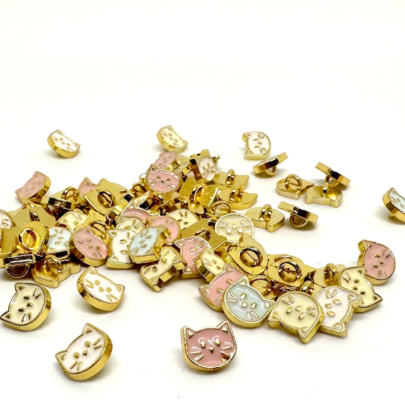 May include: A collection of gold-toned metal buttons with a cat face design. The buttons are available in three colors: pink, white, and light blue.
