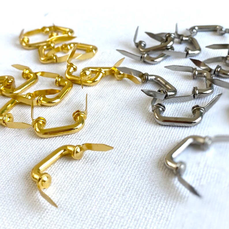 K&ouml;nnte beinhalten: Nahaufnahme von zwei S&auml;tzen Metall-Schubladengriffen. Ein Satz ist golden und der andere silbern. Die Griffe haben alle die Form einer einfachen Schleife.